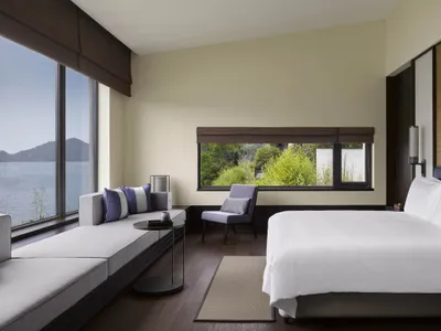 A minimalist bedroom featuring a large window with a view of lush greenery and water.