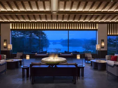 A modern hotel lounge featuring concrete and wood materials with a fireplace and seating areas by a large window overlooking