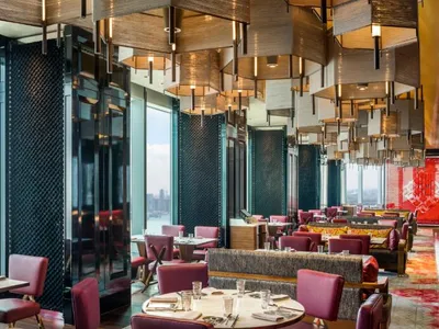 Modern restaurant interior with high ceiling, metallic hanging lights, and large windows overlooking a cityscape.