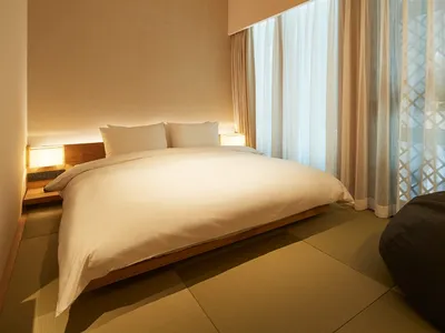 A minimalist bedroom featuring a white bed with wooden frame and fabric bedding, set against a neutral background with soft l