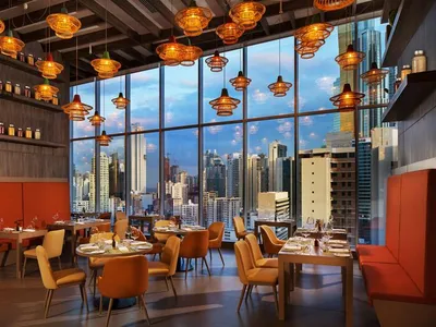 Modern restaurant interior with orange and brown lighting, large windows overlooking urban skyline, and tables set for dining