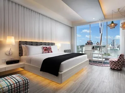 Modern hotel room with bed, desk, chairs, and large windows overlooking a cityscape.