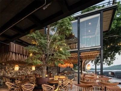Outdoor seating area of a modern restaurant with wooden tables and chairs, glass walls, and a large tree.