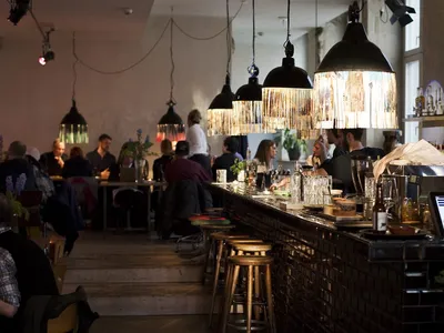 A bustling restaurant interior with industrial design elements, including metal pendant lights, glass displays, and wooden ba