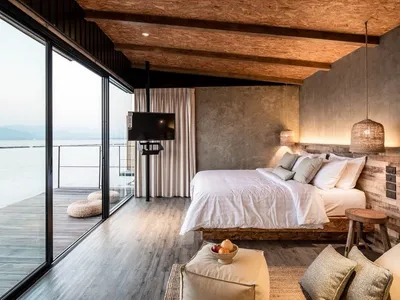 A minimalist bedroom with a white bed, wooden floor, concrete walls, and fabric accents.