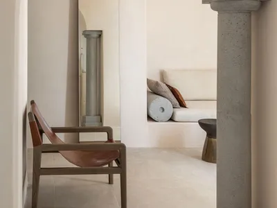 A minimalist living room with concrete walls, a brown leather chair, and a sofa with pillows.