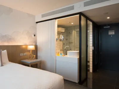 Modern minimalist hotel room with a white bed, glass door leading to a bathroom, and a wooden headboard.