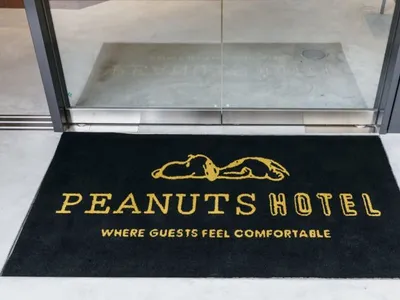 A black hotel door mat with yellow text that says 'PEANUTS HOTEL WHERE GUESTS FEEL COMFORTABLE'. The hotel logo features a sl