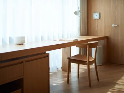 A minimalist home office space featuring wooden furniture, a window with white curtains, and a desk with a book.