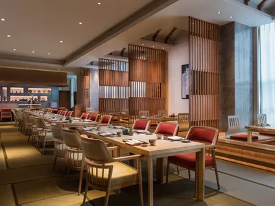 A contemporary restaurant interior featuring wooden furniture, long dining tables, red cushioned chairs, and traditional Japa
