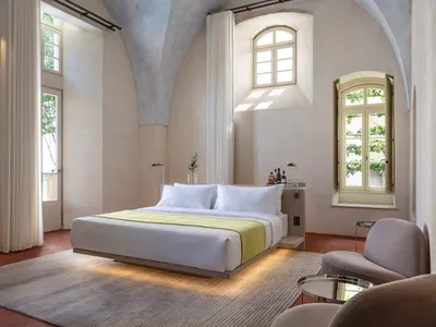 A minimalist bedroom with concrete walls and floor, featuring a large bed, a small armchair, and natural light from multiple