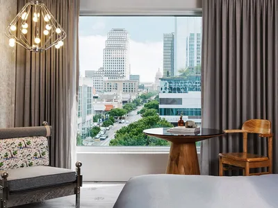 Modern minimalist hotel room with a view of the city, featuring a gray bed, a wooden chair, a round glass table, and fabric c