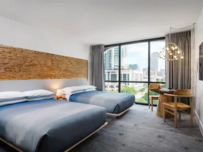 A modern hotel room with two beds, a desk, and a large window providing a view of the city.
