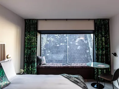 A modern minimalist bedroom featuring a bed with a wooden headboard and white linens, a small table with a black top and chai