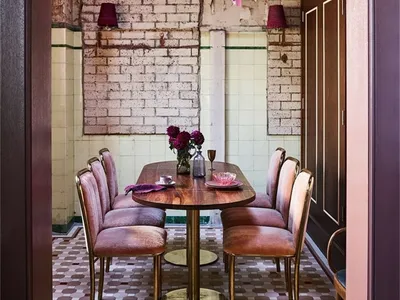 Modern restaurant dining room with metal table, velvet chairs, stone wall, and tile floor.