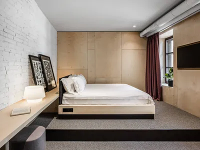 A minimalist bedroom featuring a wooden bed platform with white bedding, a wooden desk with a lamp and framed art, and a wind