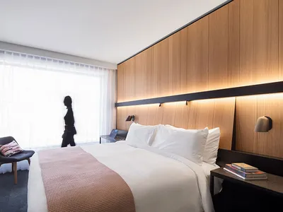 A minimalist bedroom featuring a large bed with wooden headboard, white sheets, and a side table with two books and a lamp.