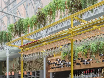 Industrial-style restaurant interior with yellow metal structures, glass hanging bottles, and wooden stools.