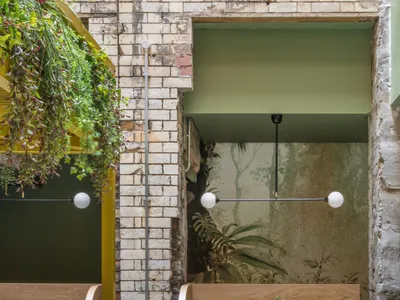 A cozy cafe interior with modern green leather seating, rattan chairs, brick walls, and hanging plants.
