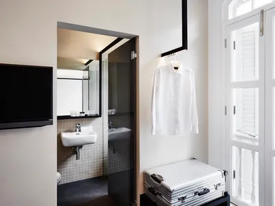 A minimalist design room in a hotel suite with a white shirt hanging on the wall, a silver suitcase, a white sink, and a tele