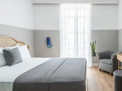 A minimalist bedroom featuring a bed with white and gray bedding, a gray armchair, a wooden desk, and a potted plant.
