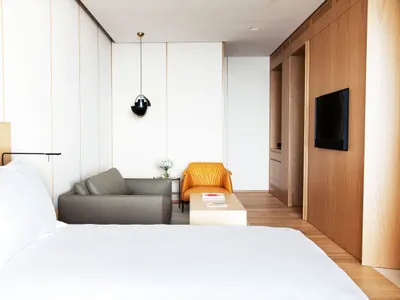 A minimalist bedroom featuring a white bed with a wooden headboard, a sofa, and an armchair, all against a background of whit