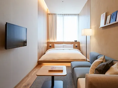 A minimalist hotel room in shades of neutral, featuring hardwood flooring and cushioned upholstered furniture.