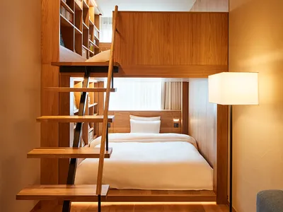 A minimalist bedroom with a wooden bunk bed and stairs, featuring a white pillow and a floor lamp.