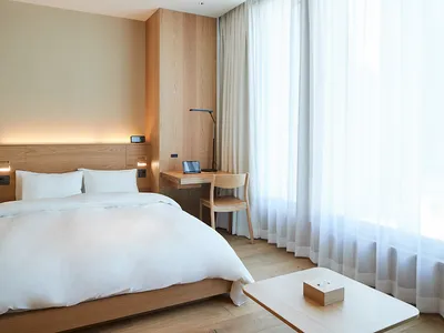 Modern minimalist hotel room with a wooden bed and glass window, featuring white textiles and a functional desk area.