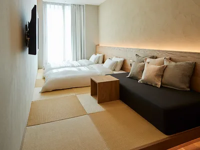 A minimalist bedroom featuring a wood-framed bed and a black sofa with cushions, set against a pale neutral background with s