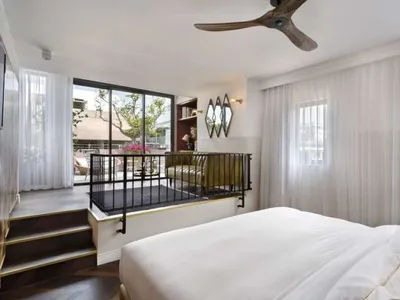 A modern bedroom featuring a glass wall with a view of the outdoors, a white bed with gold accents, and a balcony.
