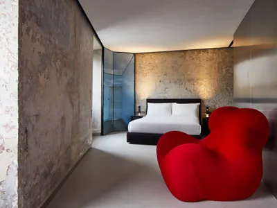 Modern bedroom featuring a concrete wall, a red chair, and a neatly made bed with white sheets and pillows.