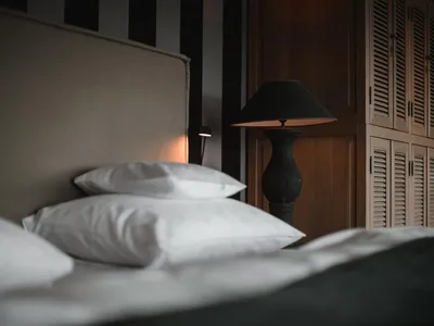 A modern bedroom with a dark headboard and white bedding. The room features a black table lamp, a wooden dresser, and a bed w