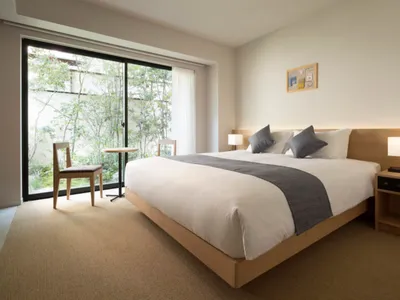 Modern minimalist bedroom with a bed, nightstands, and natural light from large windows showing greenery outside.