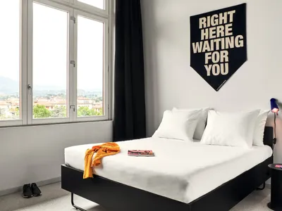 A minimalist bedroom featuring a white bed with a black headboard and a black table lamp, with a banner reading 'Right Here W