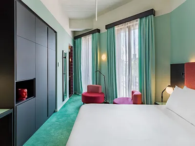 A modern hotel room with green carpet, turquoise curtains, and concrete flooring.
