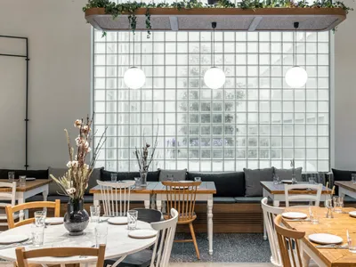 A modern cafe interior with white walls, glass block wall, wood and white tables, and bench seating.