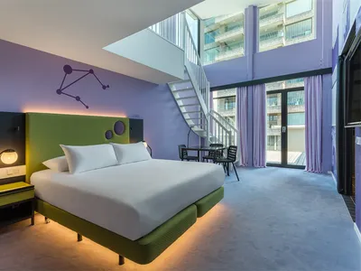A modern hotel room featuring a green and purple color scheme with a king-sized bed, green bedding, and a purple headboard.