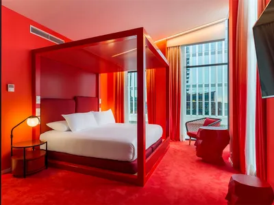 A modern hotel room with a red theme, featuring a metal frame canopy bed and red carpeting.