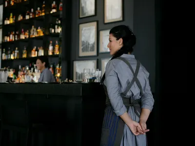 A woman wearing an apron standing at a bar with a backdrop of shelves filled with bottles and framed pictures on the wall.