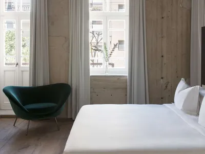 A minimalist bedroom featuring a white bed with white linens, a green armchair, and white curtains.