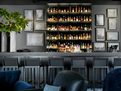 Modern bar interior with sleek metallic shelving, glass bottles, leather seating, stone countertops, and wooden accents.