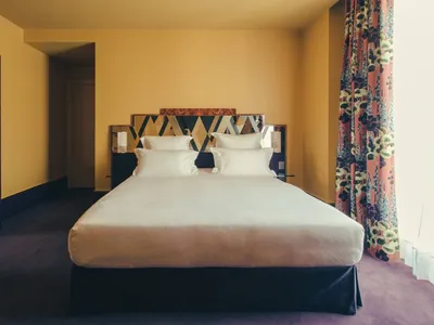 A spacious modern bedroom featuring a bed with white linens, a geometric headboard, and colorful curtains against a mustard y