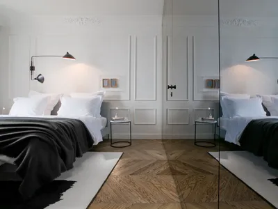 A minimalist bedroom with a wood floor and white walls, featuring a bed with white bedding and a dark throw blanket, illumina