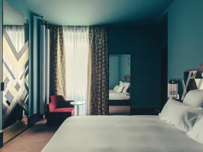 A contemporary hotel room with textured and patterned wallpaper, a bed with white bedding, and a red armchair.