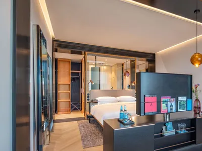 Modern hotel room with wooden flooring and glass walls, featuring a bed, nightstand, and magazines.
