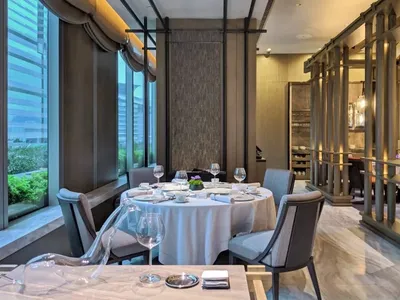Modern restaurant dining room with marble flooring, metal railings, and floor-to-ceiling windows overlooking cityscape.