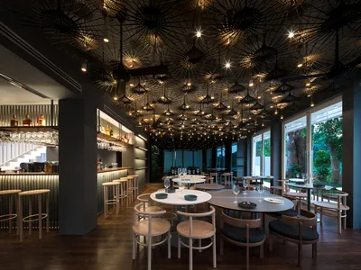 A modern restaurant interior with paper umbrellas hanging from the ceiling, wooden furniture, and a bar area with liquor bott