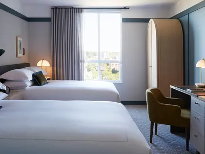 A modern hotel room with two double beds, a desk, and a window overlooking trees.