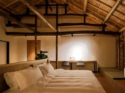 A minimalist bedroom featuring wooden beams on the ceiling, a bed with white bedding, and a small desk area.